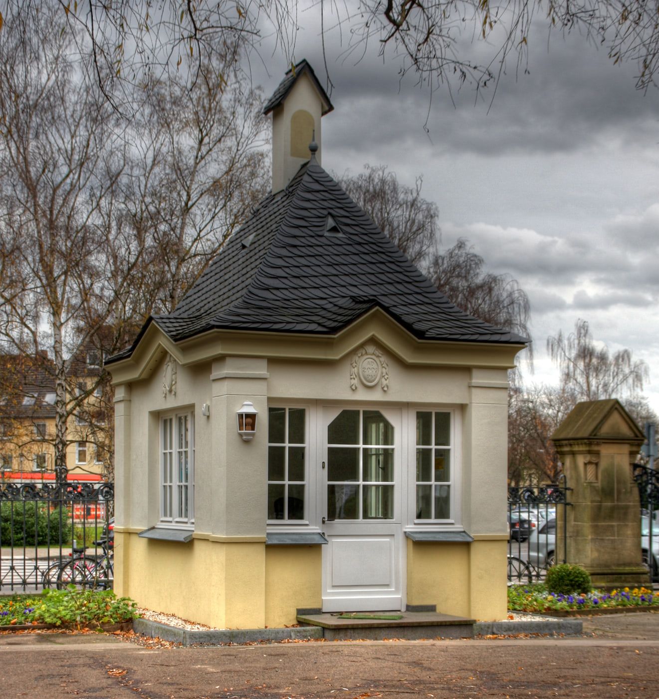 Kleines hellgelbes Pförtnerhaus mit steilem polygonalem Schieferdach, weiß gerahmten Fenstern und schlankem Dachaufsatz unter kahlen Birken auf Südfriedhof Köln
