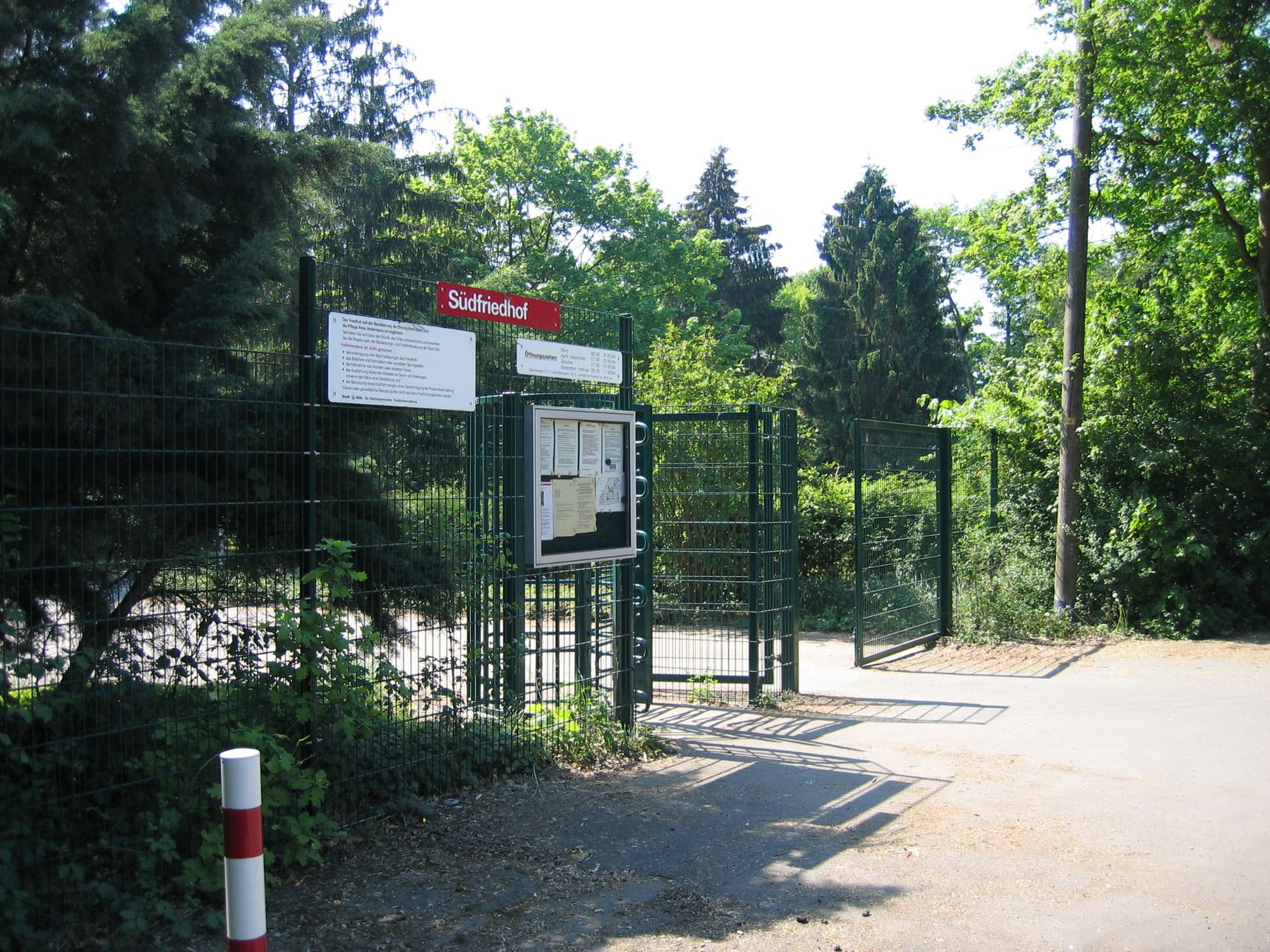 Geöffneter Seiteneingang mit grünen Gitterzäunen, rotem Südfriedhof-Schild und Informationskasten zwischen dichtem Grün auf Südfriedhof Köln