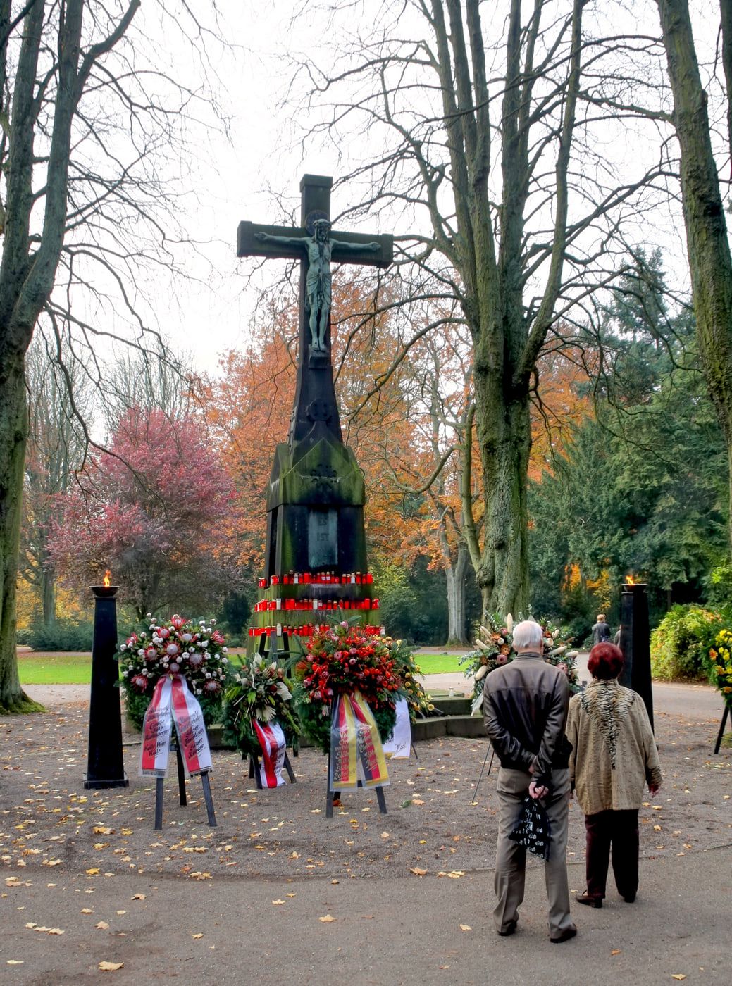 Das Hochkreuz am Hauptweg zu Allerheiligen, umgeben von roten Grablichtern, Kränzen, brennenden Fackeln und zwei Besuchern auf Südfriedhof Köln