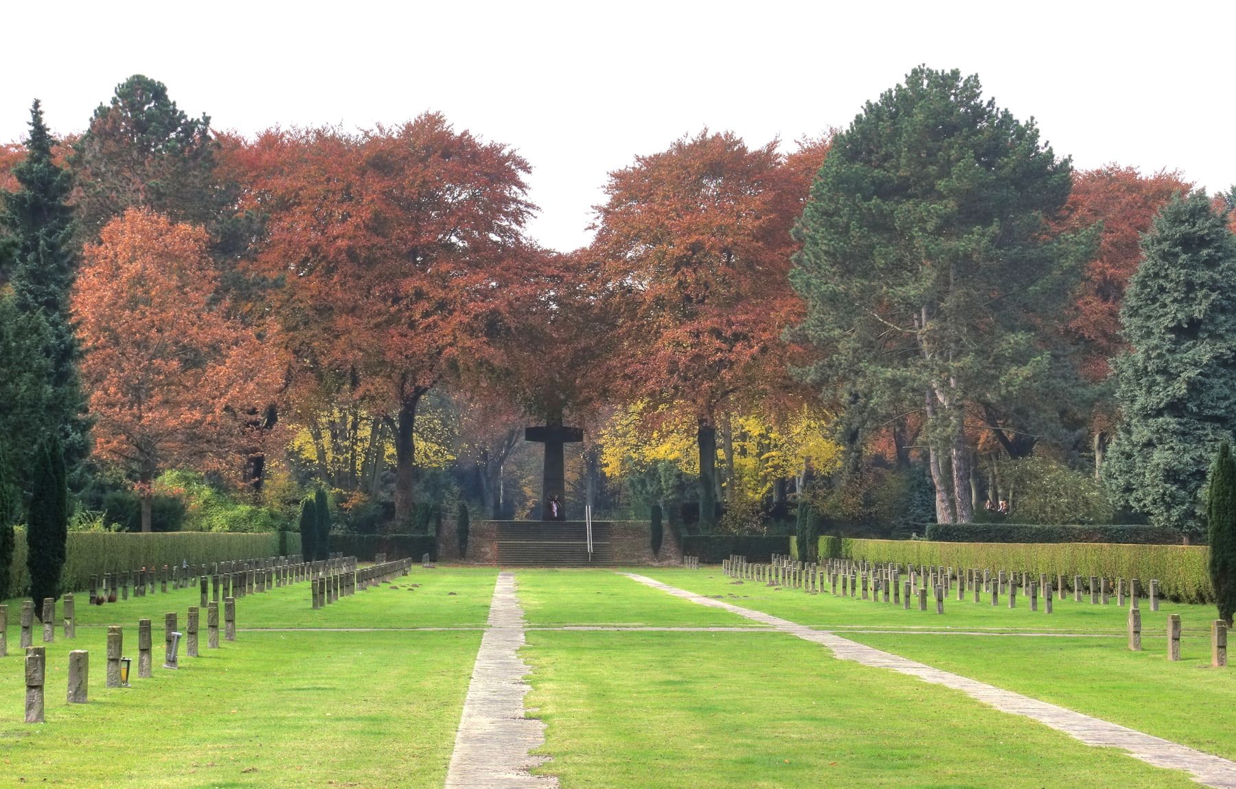 Symmetrisches Kriegsgräberfeld mit niedrigen Steinkreuzen, zwei schmalen Plattenwegen und dem südwestlichen Hochkreuz vor herbstlichen Bäumen auf Südfriedhof Köln